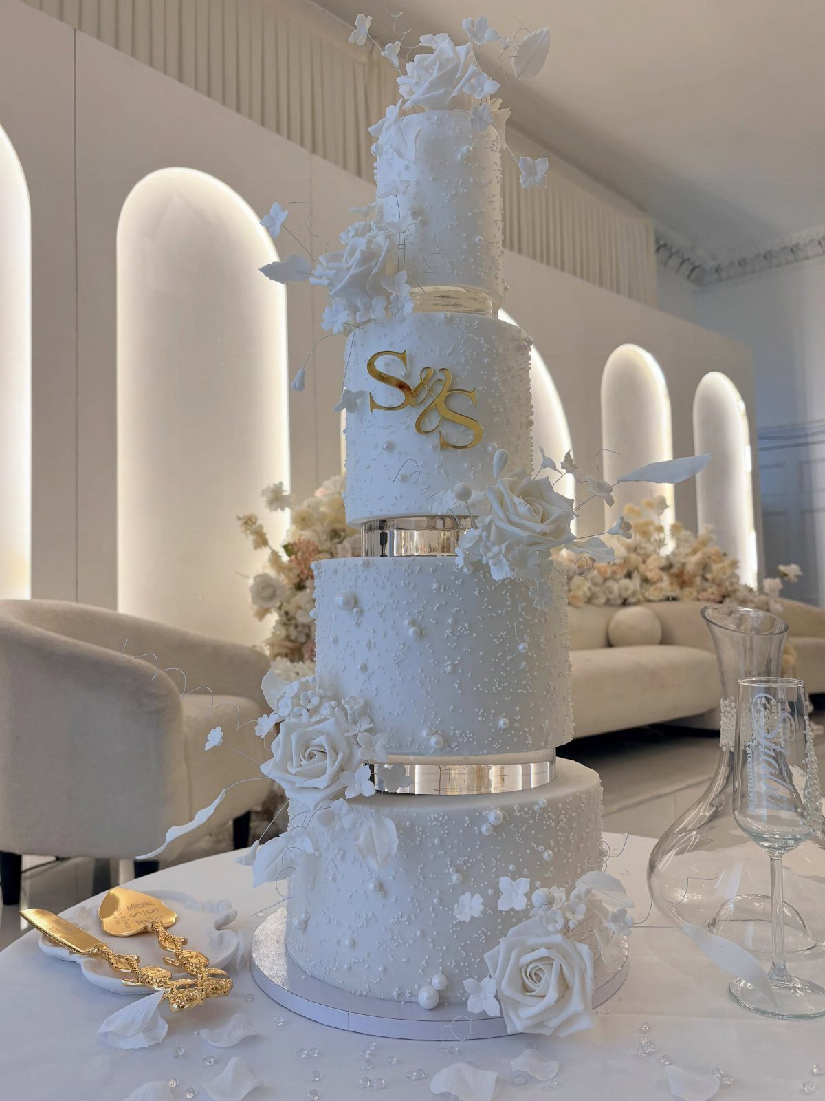 Wedding Cake Photo Ideas: 6 Designs for Every Style 5 Four-tier white wedding cake with white roses, pearls, and gold initials on a table.