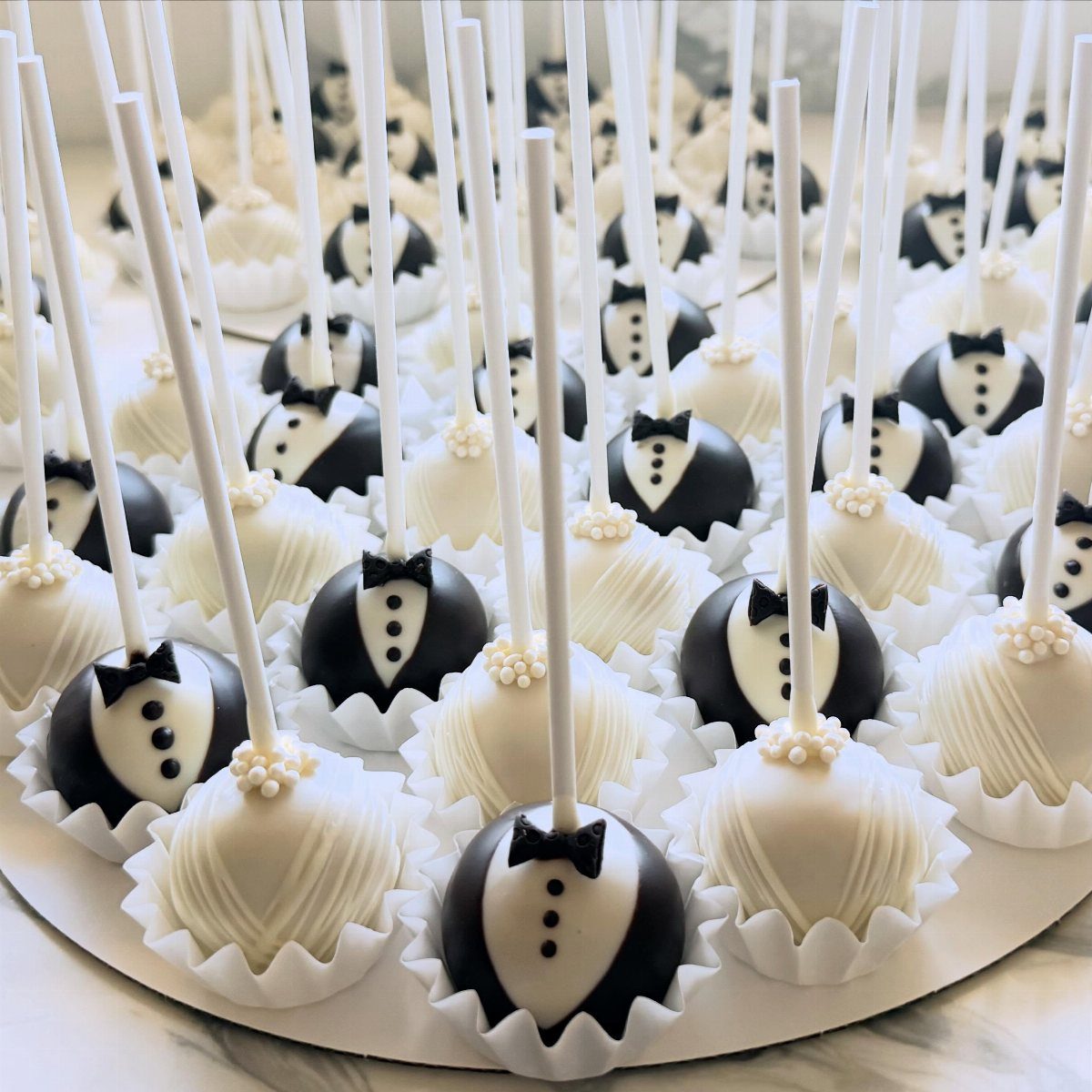 5 Wedding Cake Pops Ideas for Favors and Dessert Tables 1 Tuxedo and wedding gown cake pops arranged on a white circular platter.