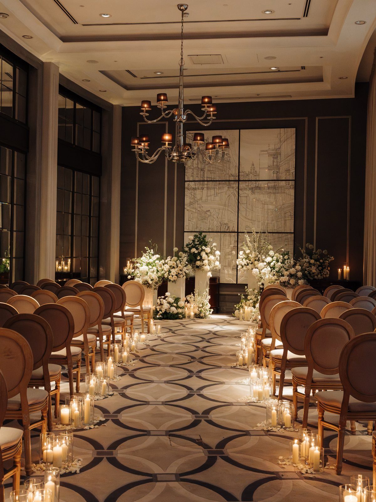 Wedding ceremony aisle with round-back chairs, white flowers, and glowing candles indoors.