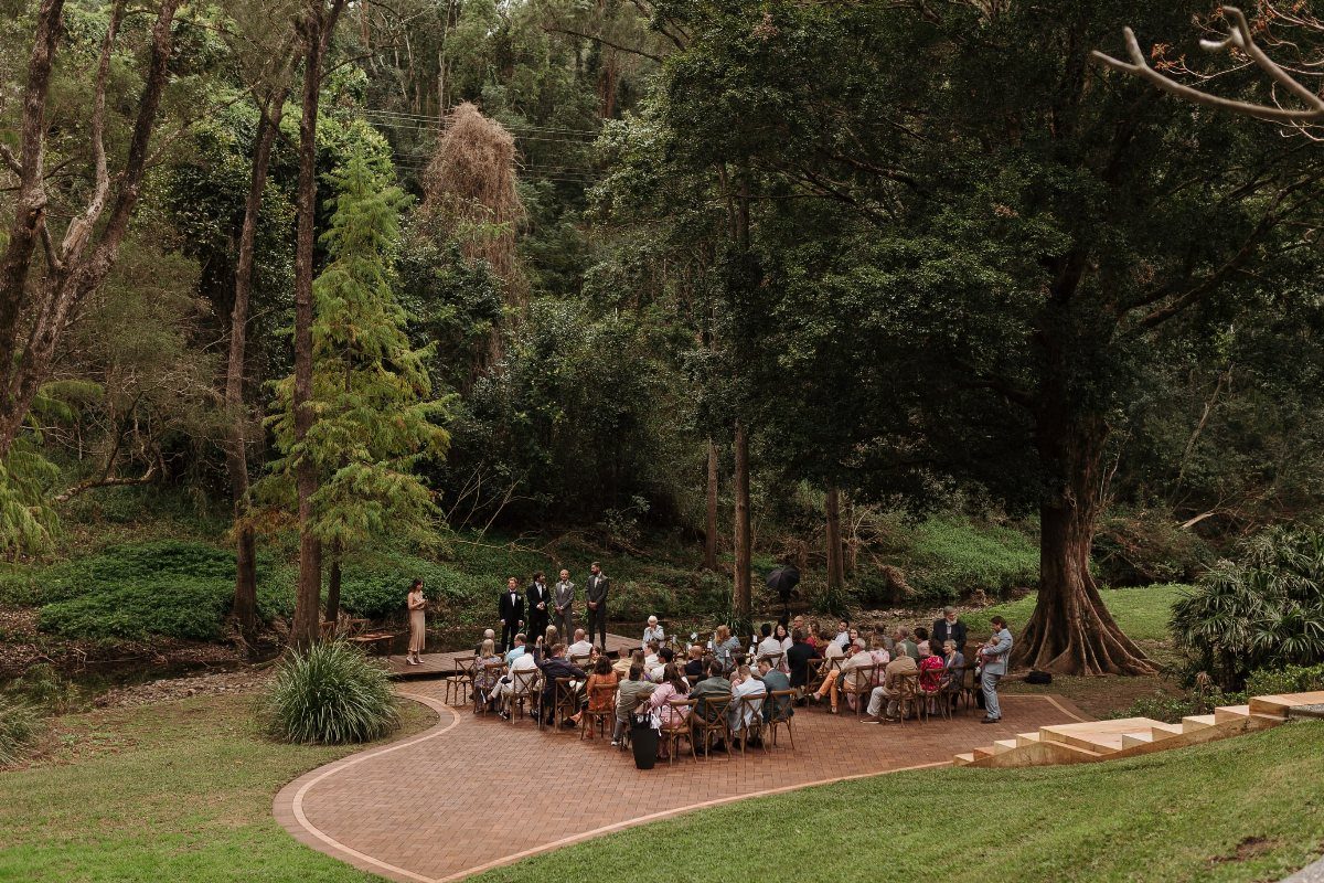 Outdoor wedding ceremony with guests seated in chairs within a dense green forest.