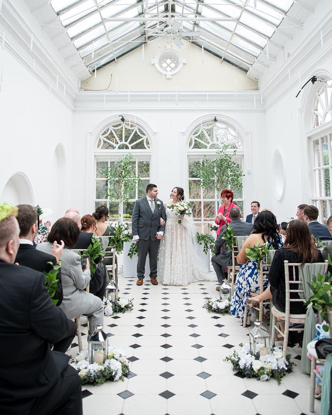 Bride and groom standing in a bright white conservatory with guests seated around.