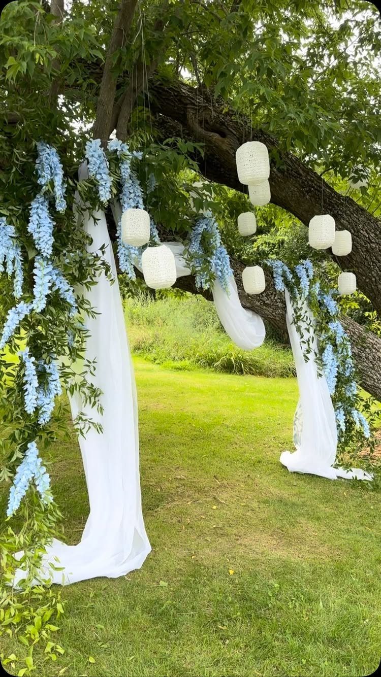 19 Wedding Decorations at Home: DIY Ideas to Try 18 White fabric drapes and blue flowers hanging from a tree with paper lanterns.
