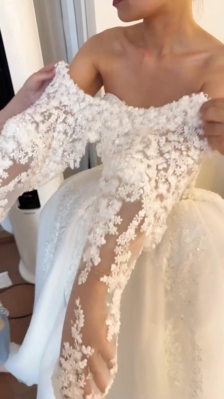 White wedding gown with intricate floral lace embroidery and sheer off-the-shoulder sleeves.
