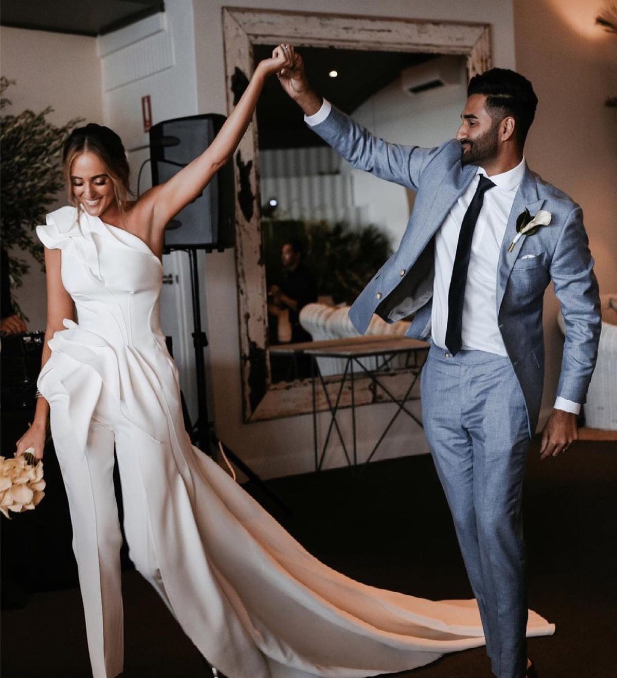 Woman in white bridal jumpsuit with train dancing with man in blue suit.