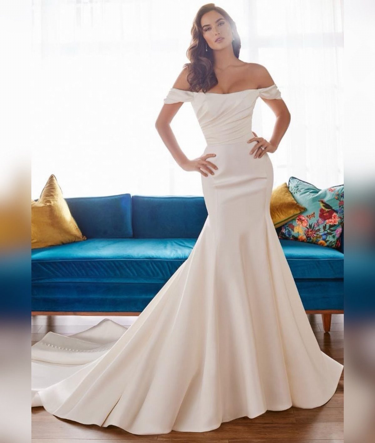 Wedding Dresses Elegant and Classy: 14 Gown Styles 13 Woman in a white off-the-shoulder satin trumpet wedding gown posing by a blue sofa.