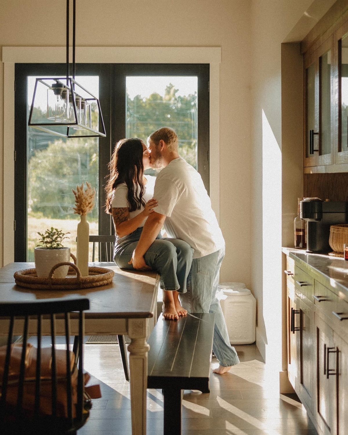 Couple kissing in a bright kitchen with the woman sitting on a bench.