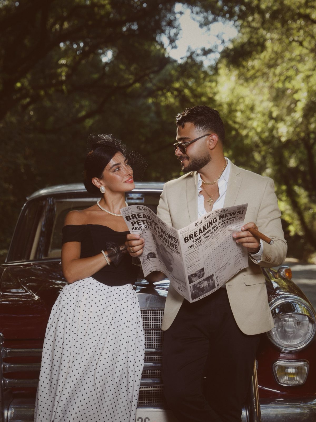 Couple in vintage attire leaning against a classic red car in a forest.