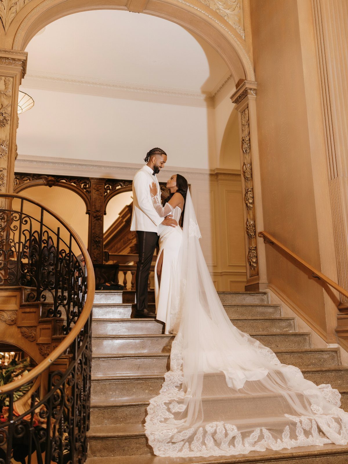 Wedding couple posing on a grand staircase under an ornate architectural archway.