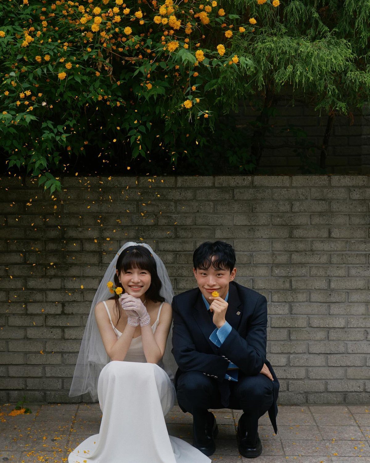 Bride and groom smiling at the camera while holding yellow flowers against a brick wall.