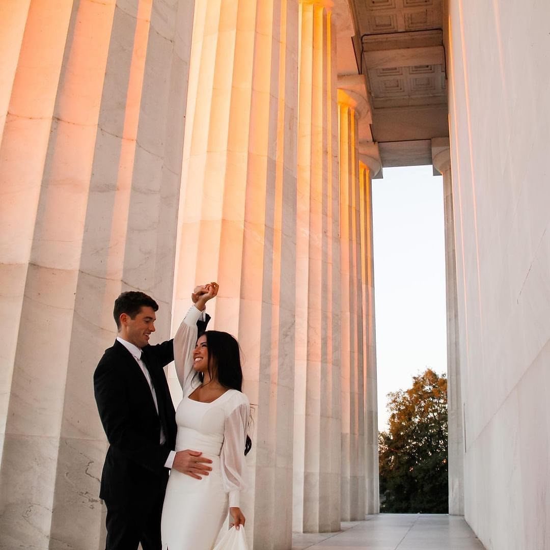 Couple dancing in white dresses and black suits between large white architectural columns.