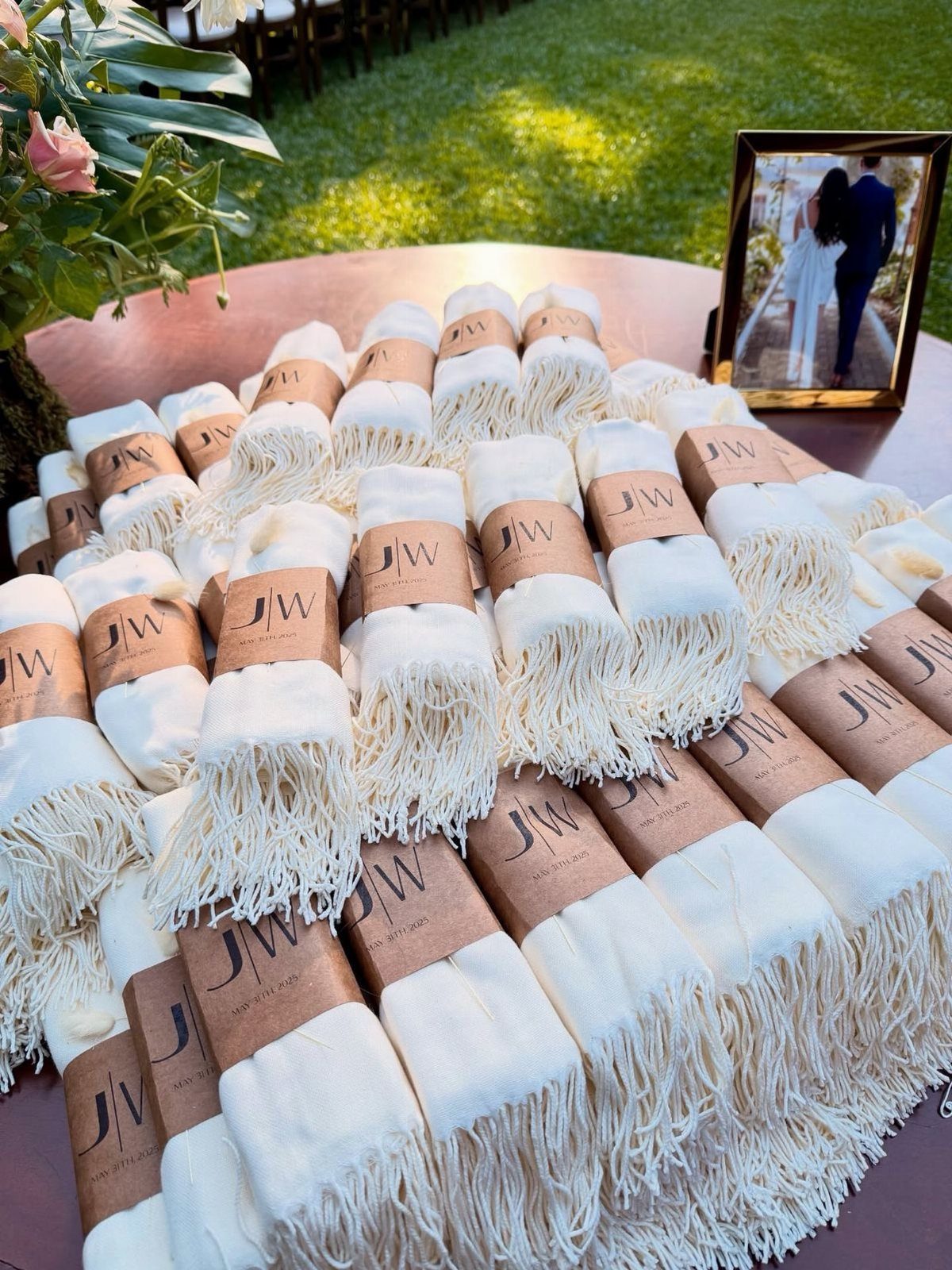 30 Wedding Guest Keepsakes Your Guests Will Actually Use 30 White pashmina shawls with brown paper bands arranged on a table outdoors.