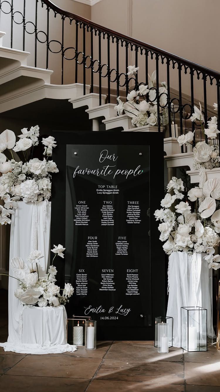 Black acrylic wedding seating chart surrounded by white flowers and white fabric draping.
