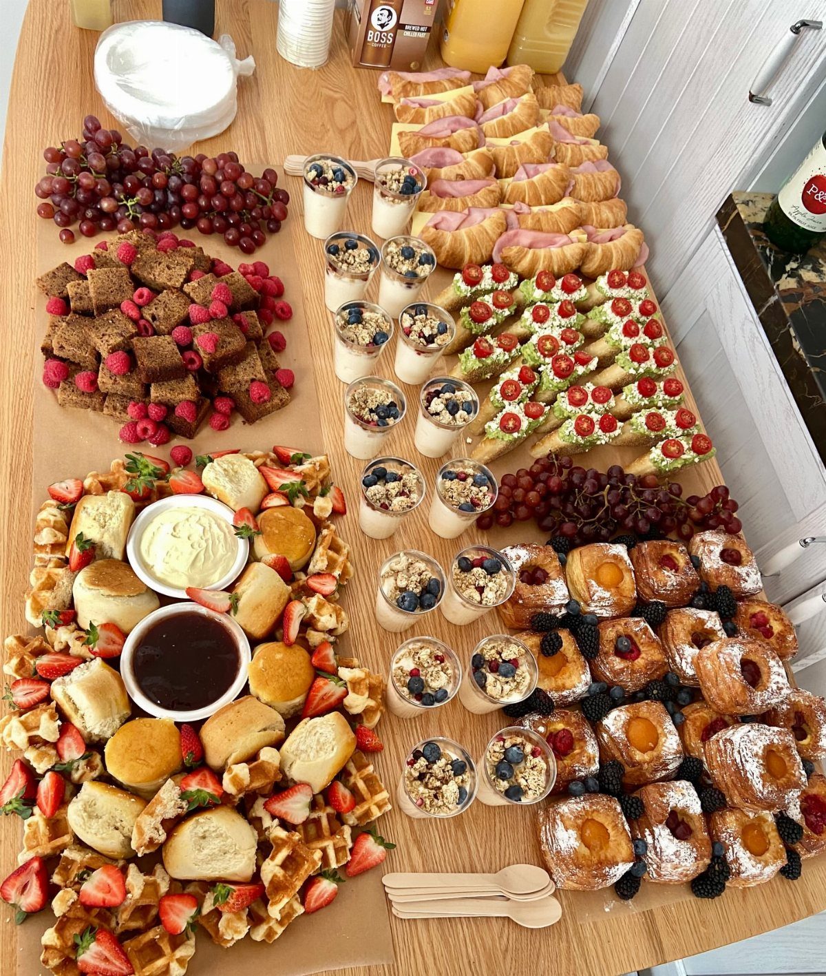 Brunch food spread with waffles, pastries, fruit parfaits, and berries on a wooden table.