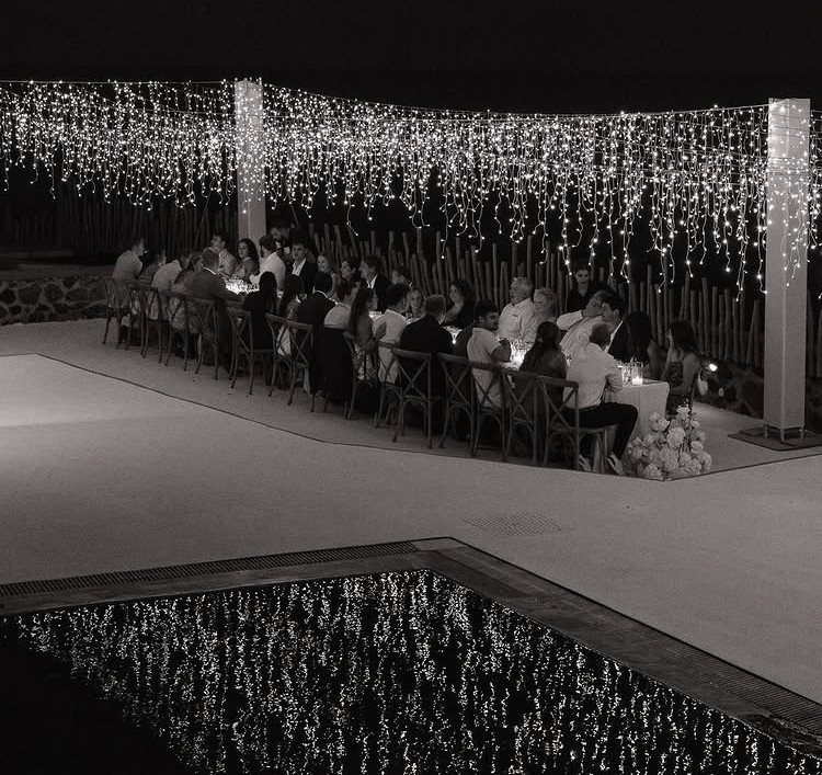 Outdoor wedding dinner table with guests seated under a canopy of twinkling fairy lights.