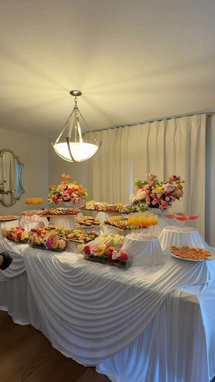 Buffet table with white draped cloth, assorted food platters, and colorful flower arrangements.