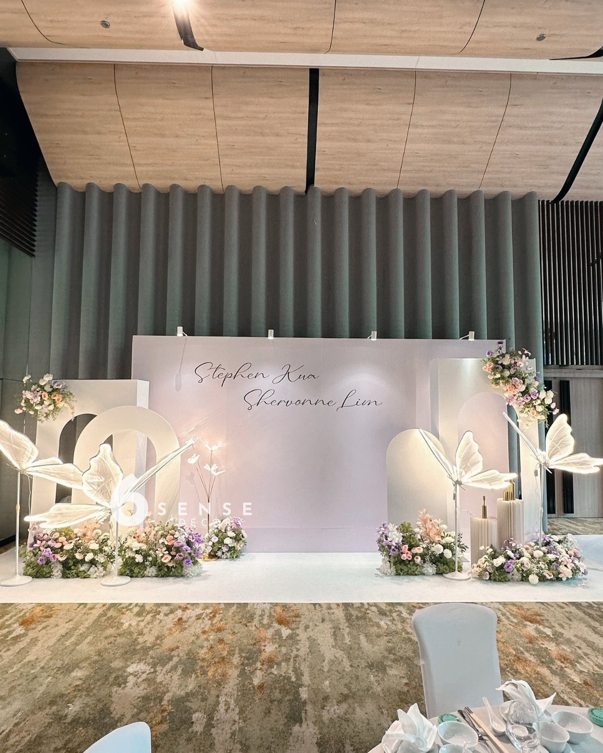 White wedding backdrop with cursive names, large butterfly accents, and pastel floral arrangements.