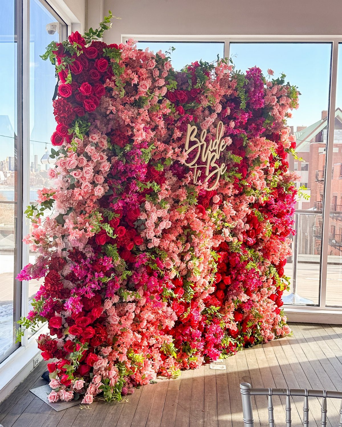 Flower wall with pink and red blooms and a gold Bride to Be sign.
