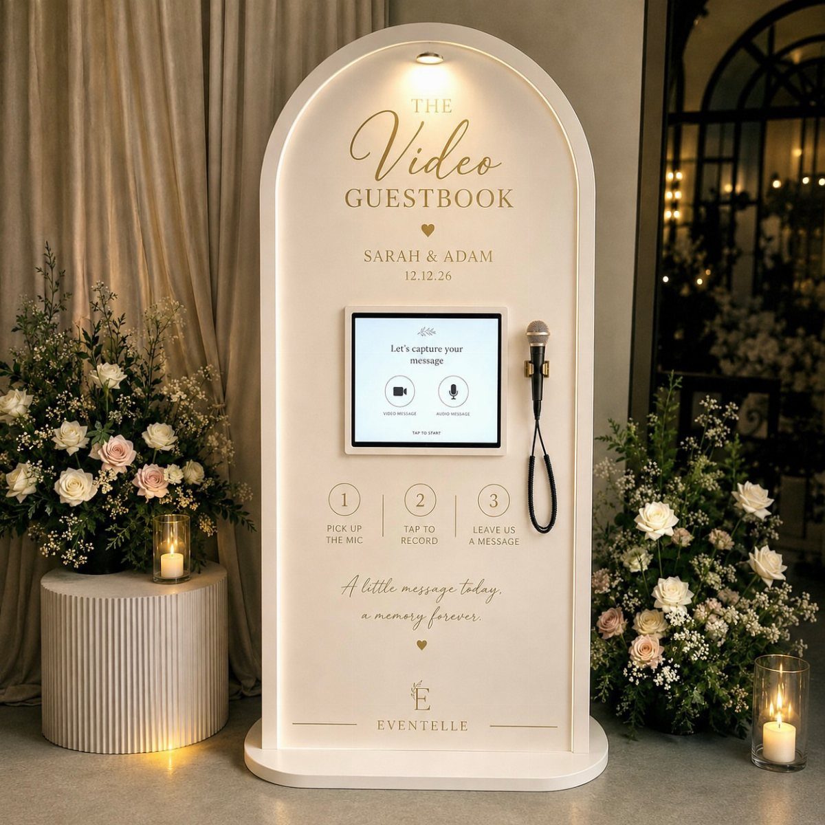 White arched video guestbook kiosk with microphone and screen surrounded by floral arrangements.