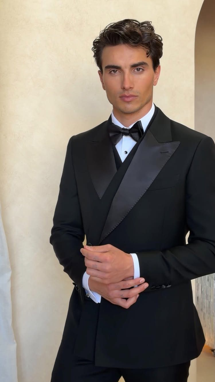 Man in a black tuxedo with a bow tie posing against a beige wall.