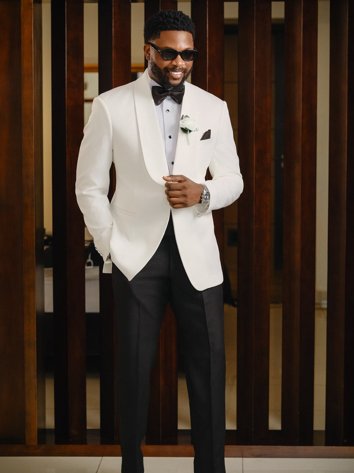 Man in a white tuxedo jacket and black trousers posing against wooden slats.