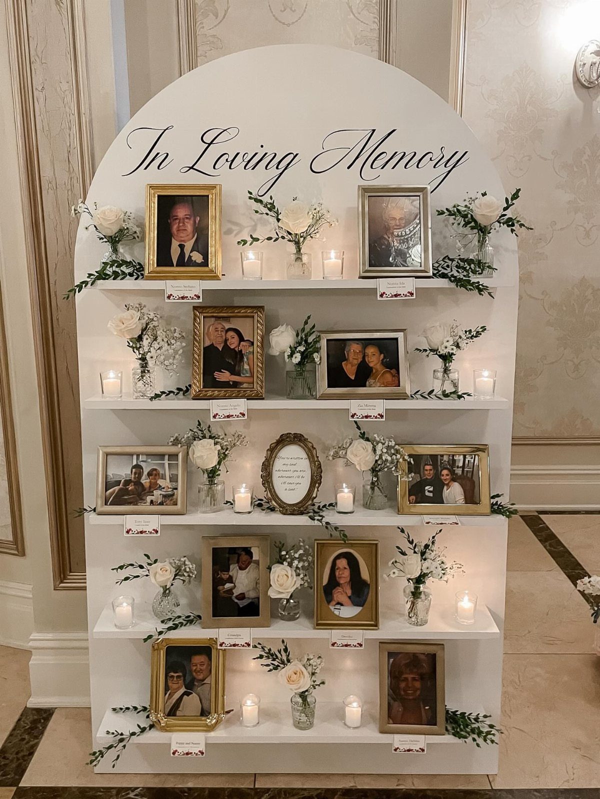Memorial display with framed photos, white flowers, and candles on a white arched shelf.