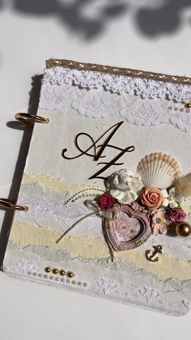 Decorated scrapbook cover with lace, gold initials, seashells, and small fabric flowers.