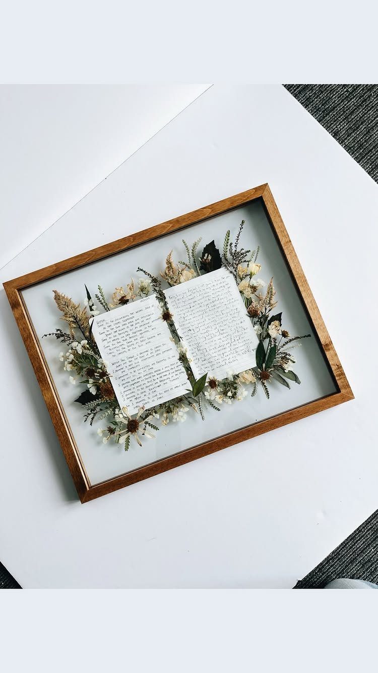Open handwritten vows booklet surrounded by dried flowers inside a wooden shadow box frame.