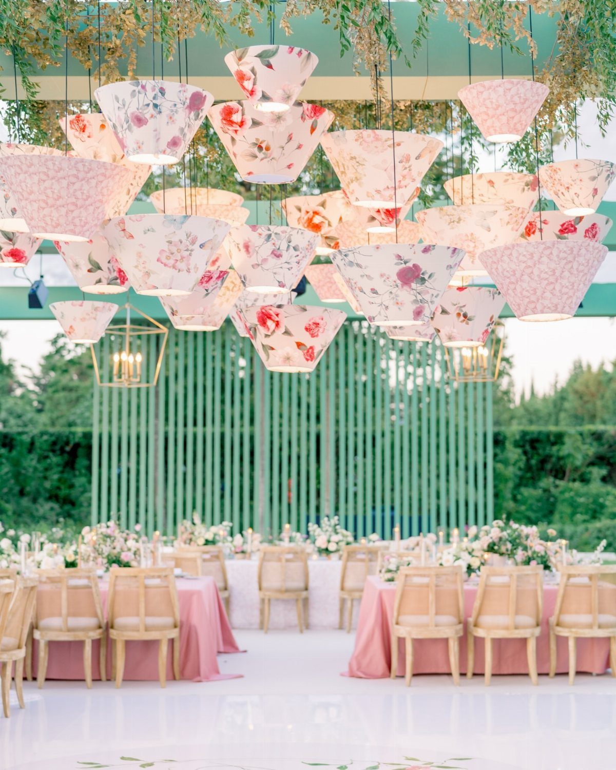 Floral conical lamps hanging from a green pergola over wedding reception tables.