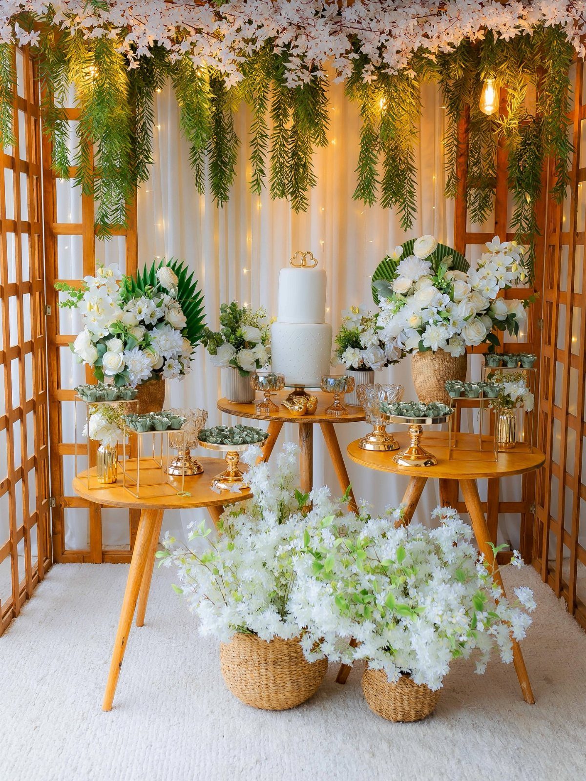 Wedding cake and white flowers under a wooden pergola with hanging greenery and lights.