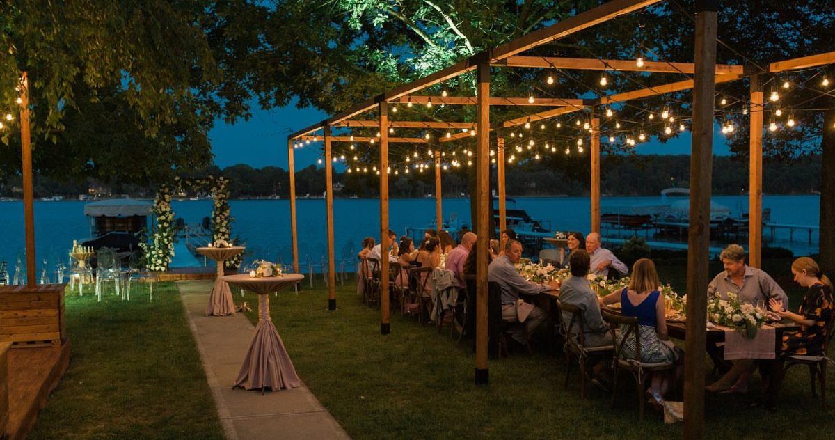 Wooden pergola with string lights over a long dining table by a lake