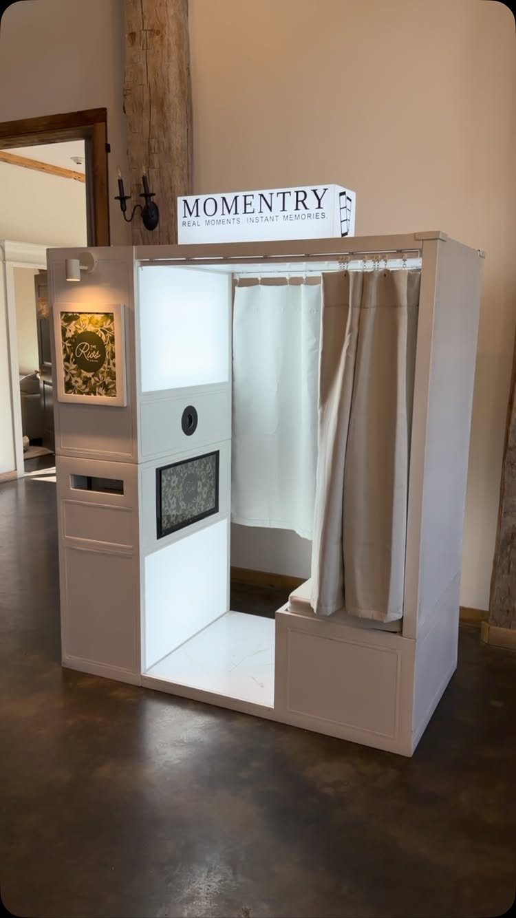 Best Wedding Photo Station Ideas: 8 Booths and Backdrops 1 White enclosed photo booth with a curtain and Momentry sign in an indoor setting.