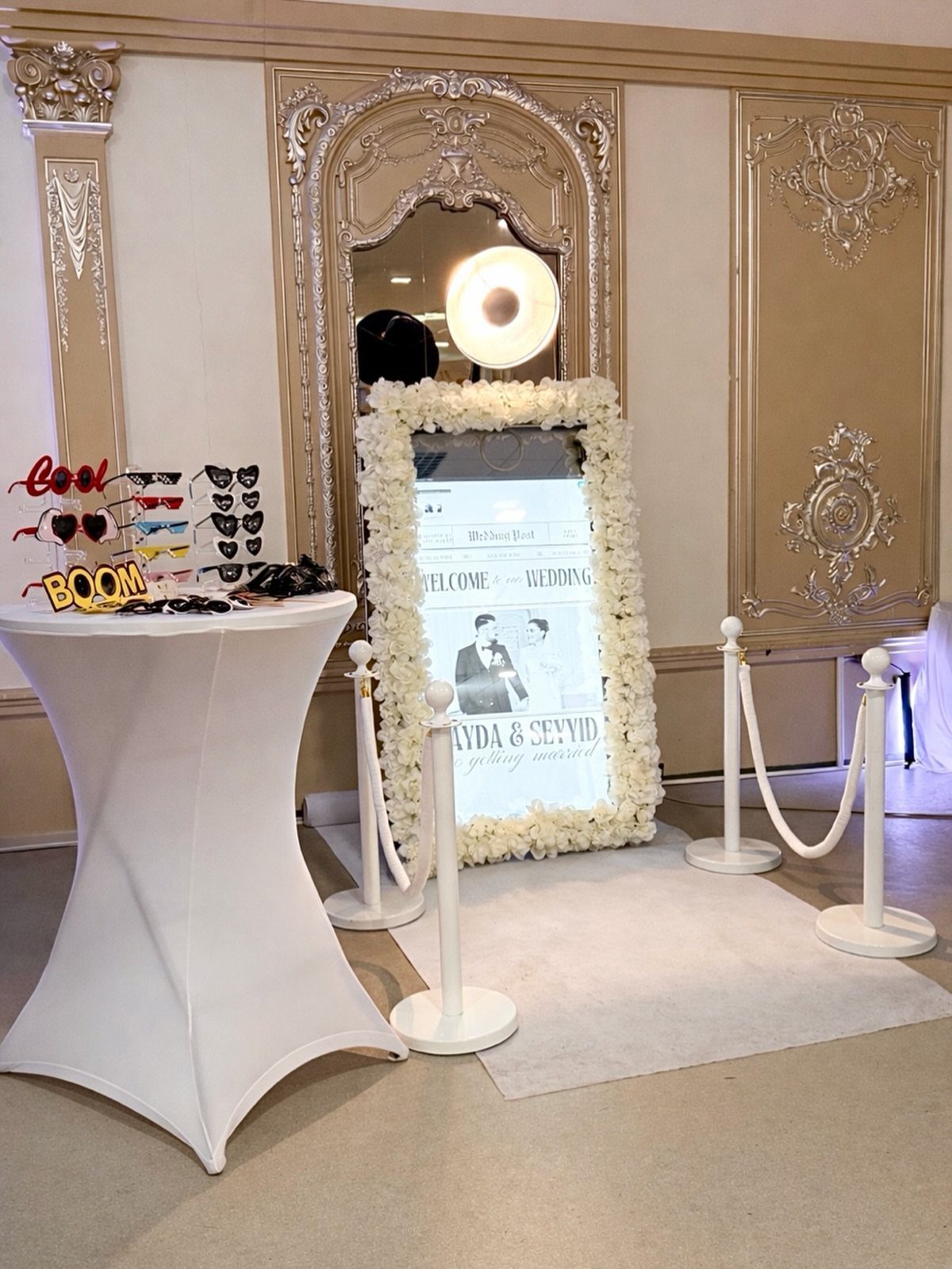 Best Wedding Photo Station Ideas: 8 Booths and Backdrops 3 Mirror photo booth with floral frame and prop table at a wedding venue.