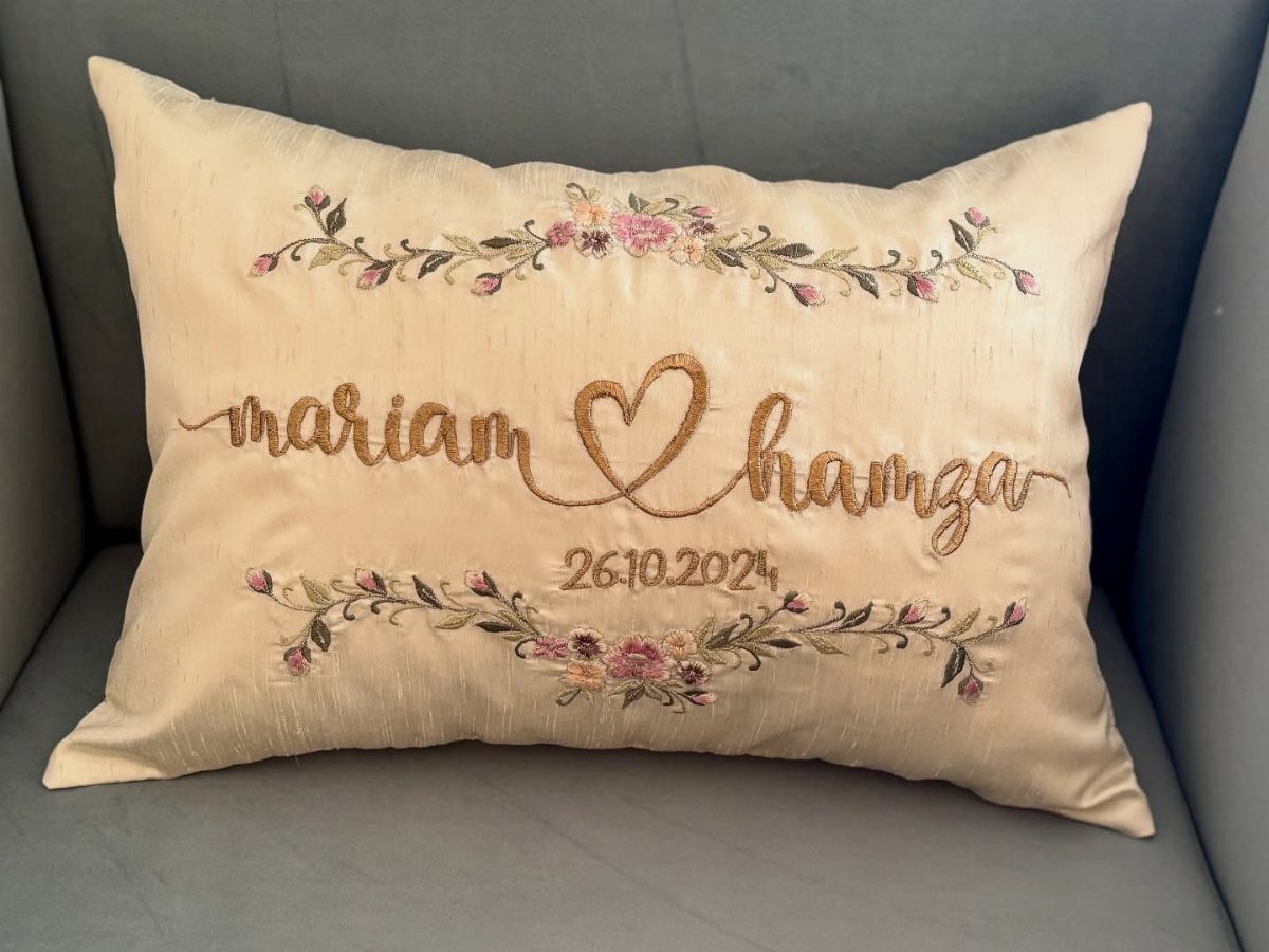 22 Wedding Present Ideas for Couples to Treasure 3 Cream pillow with gold embroidered names, a heart, and floral patterns on grey fabric.