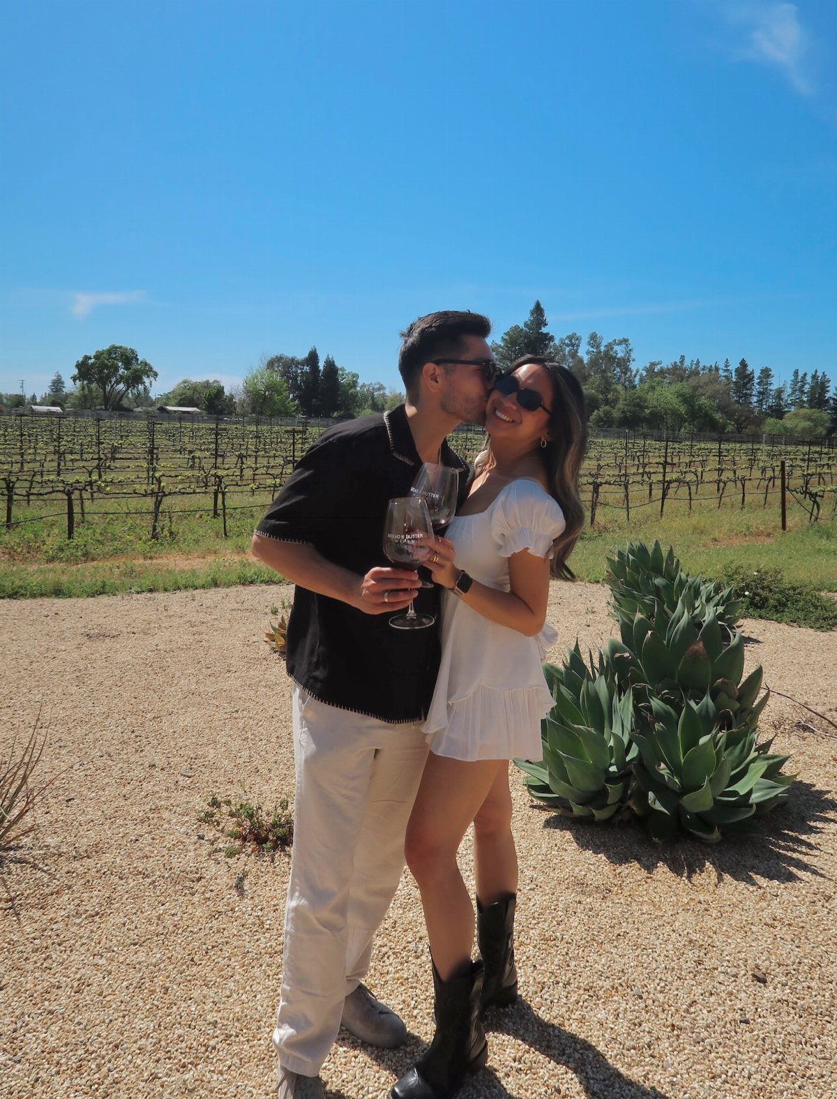 22 Wedding Present Ideas for Couples to Treasure 1 Couple holding glasses of red wine while kissing in a sunny vineyard setting.