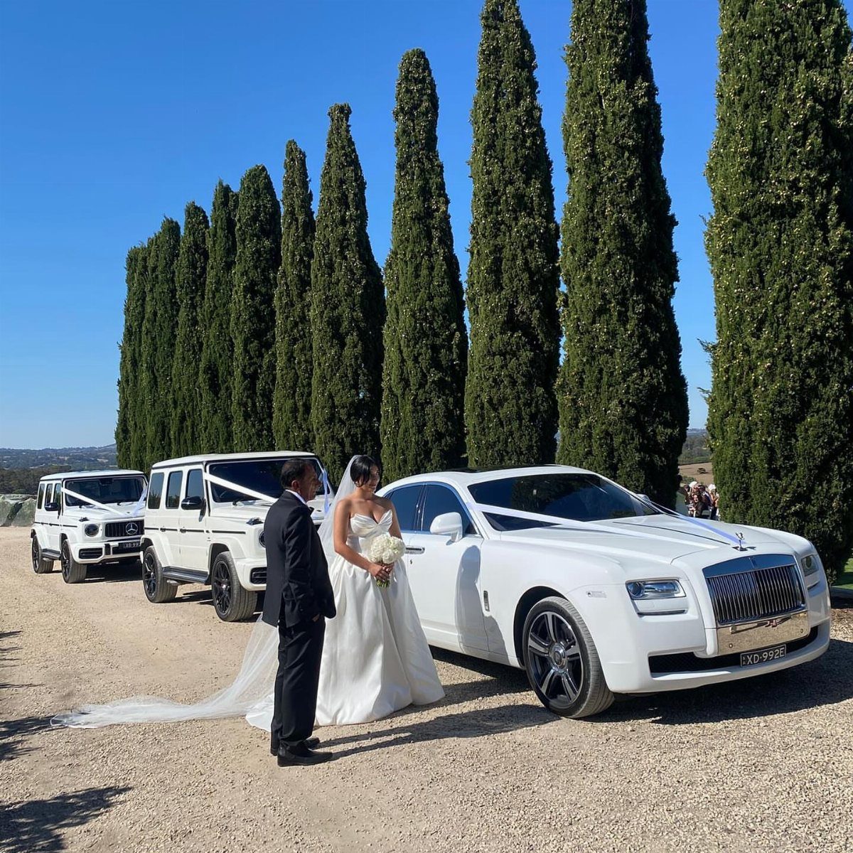 Bride and groom standing next to a white luxury car and cypress trees.