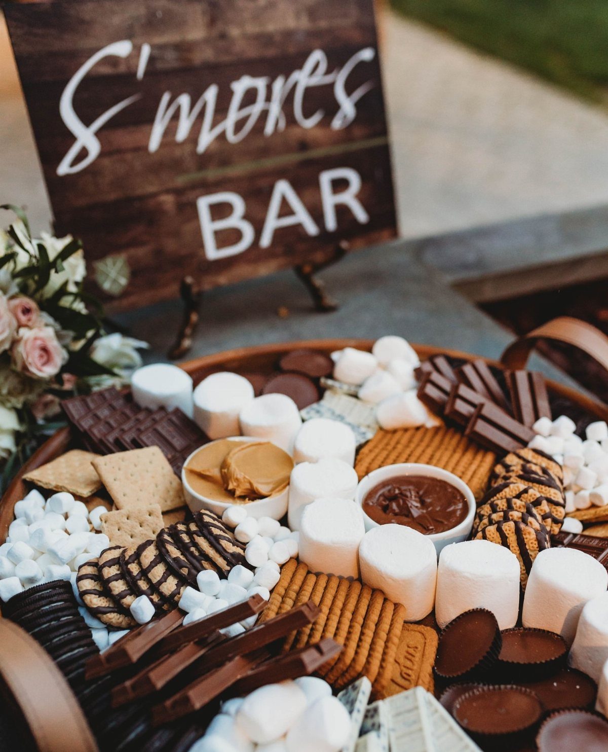 13 Wedding Reception Ideas for Guests Who Love Fun 13 S'mores bar platter with marshmallows, chocolate, and graham crackers under a wooden sign.