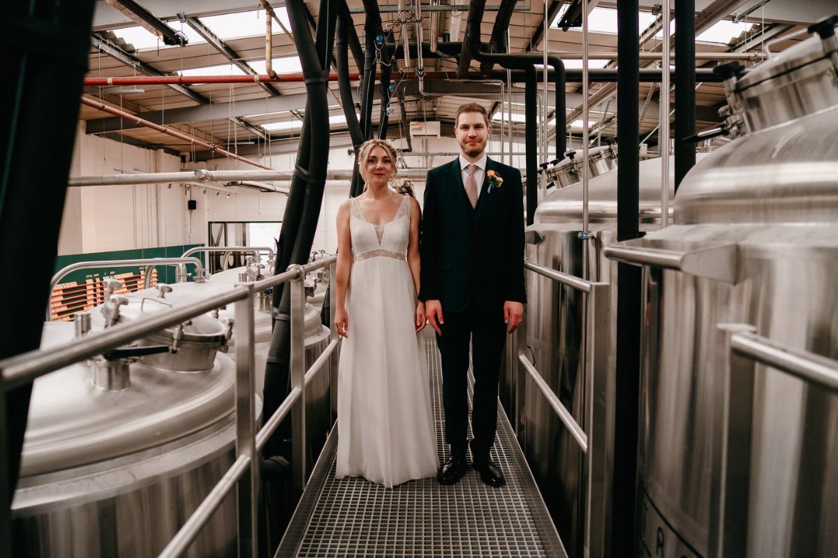 17 Wedding Reception Venues for Every Style and Budget 4 Bride and groom posing between large stainless steel tanks in a craft brewery.