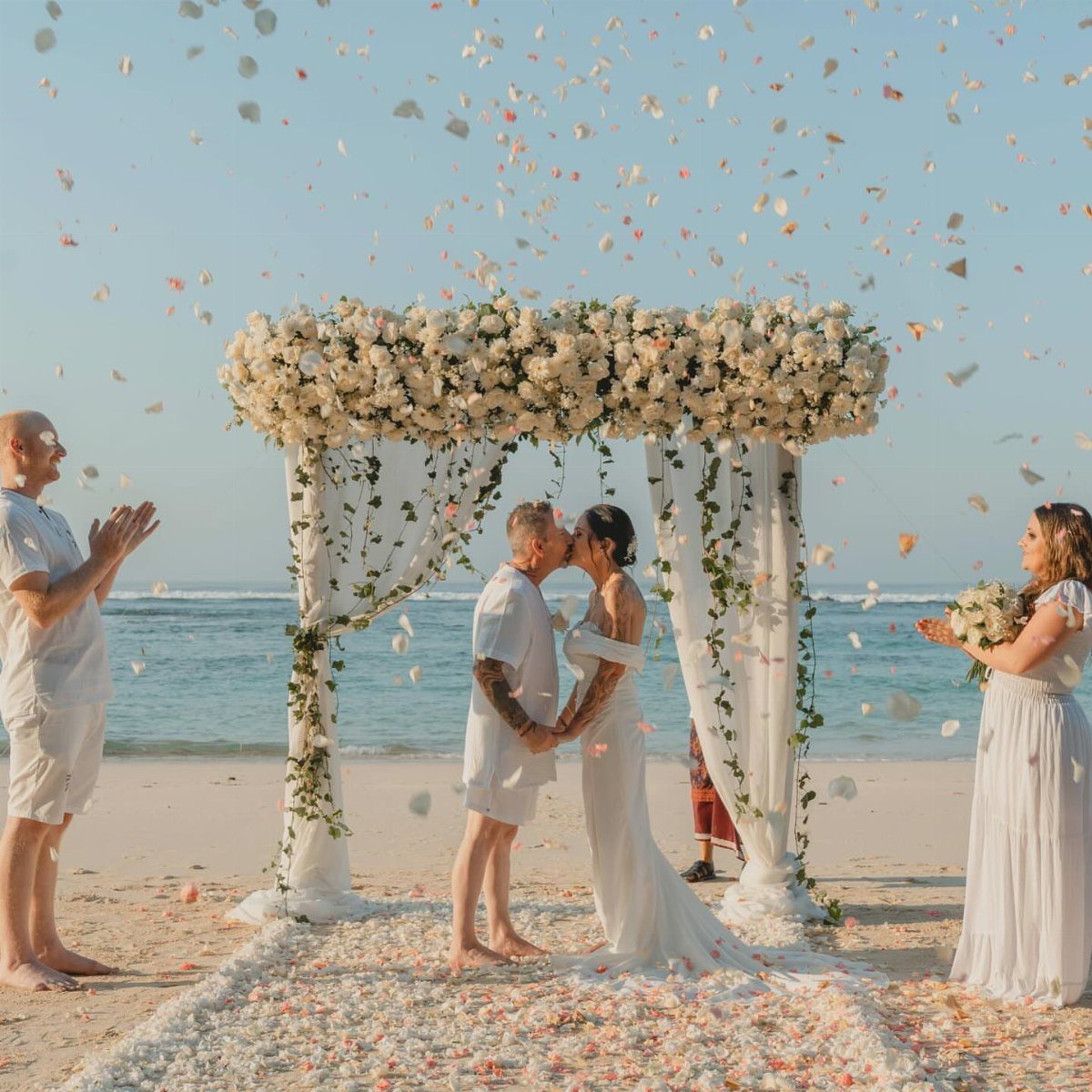 Couple kissing under a white floral arch on a beach with falling petals.