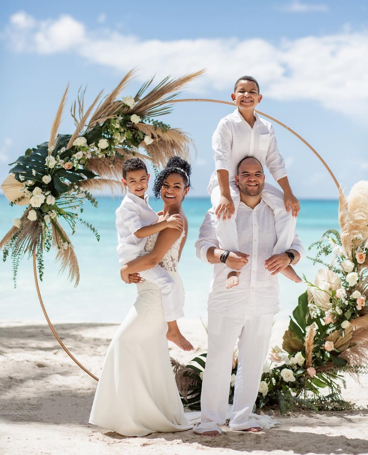 Family posing in white attire by a floral arch on a sunny beach.