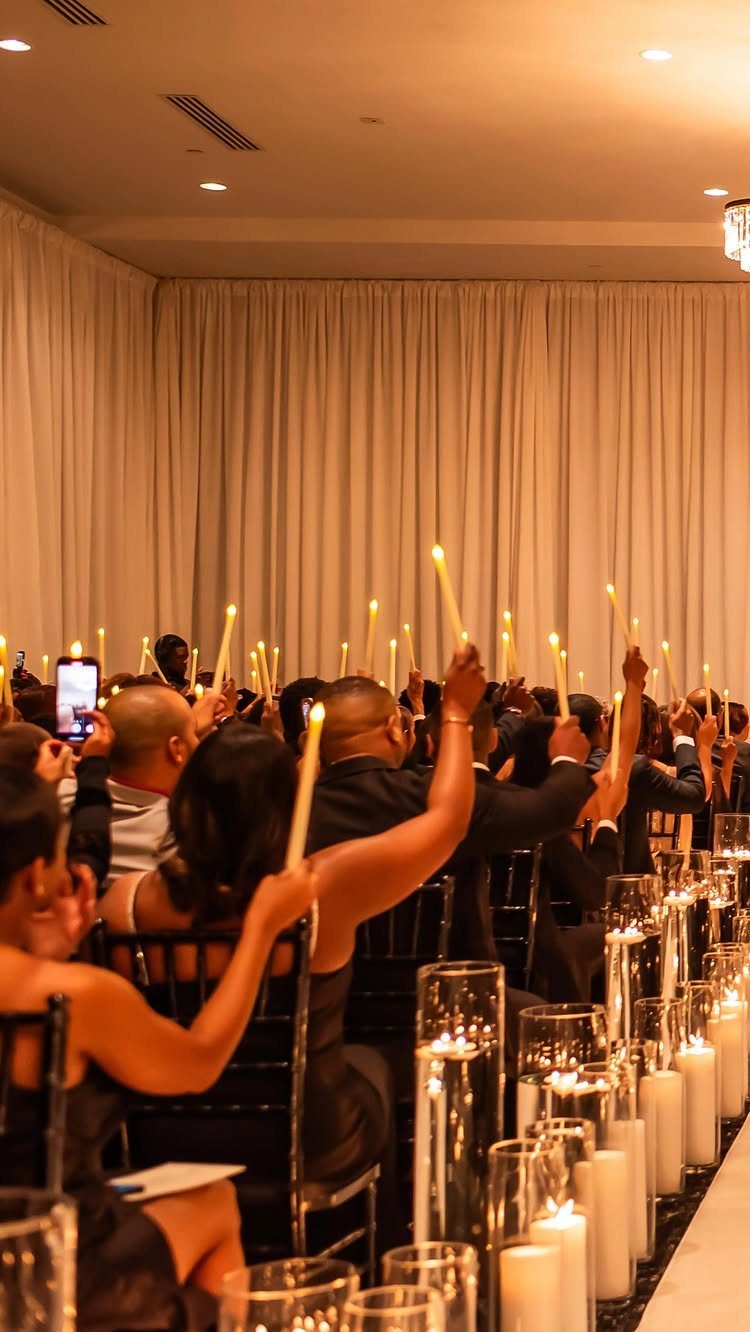 People in formal wear holding lit white candles in a room with curtains.