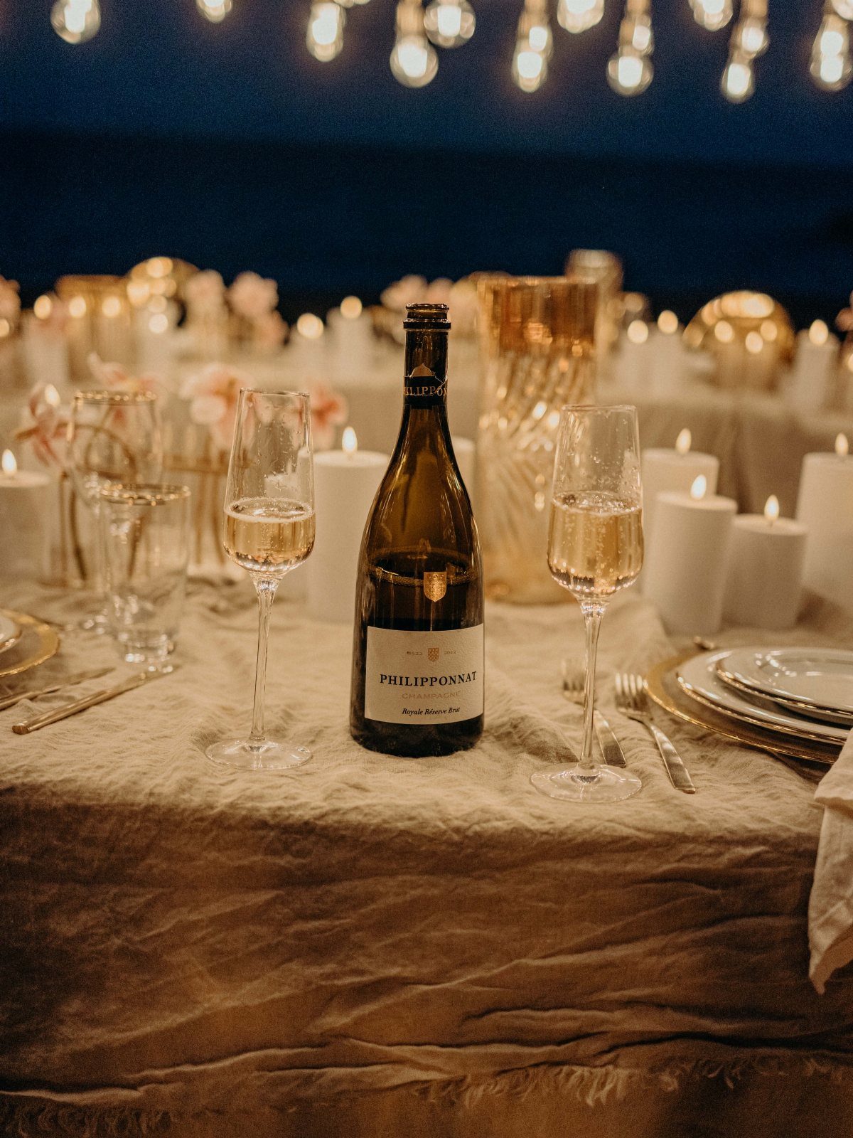 Champagne bottle and glasses on a candlelit dinner table with warm string lights.