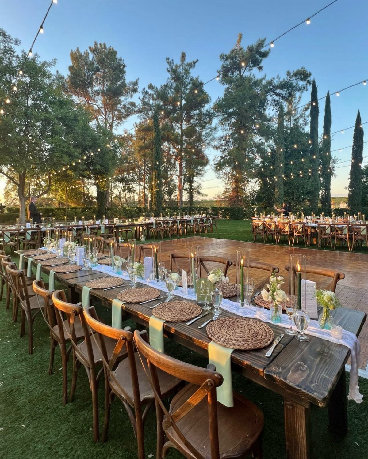 Long wooden farmhouse tables set for a wedding on a lawn with string lights.