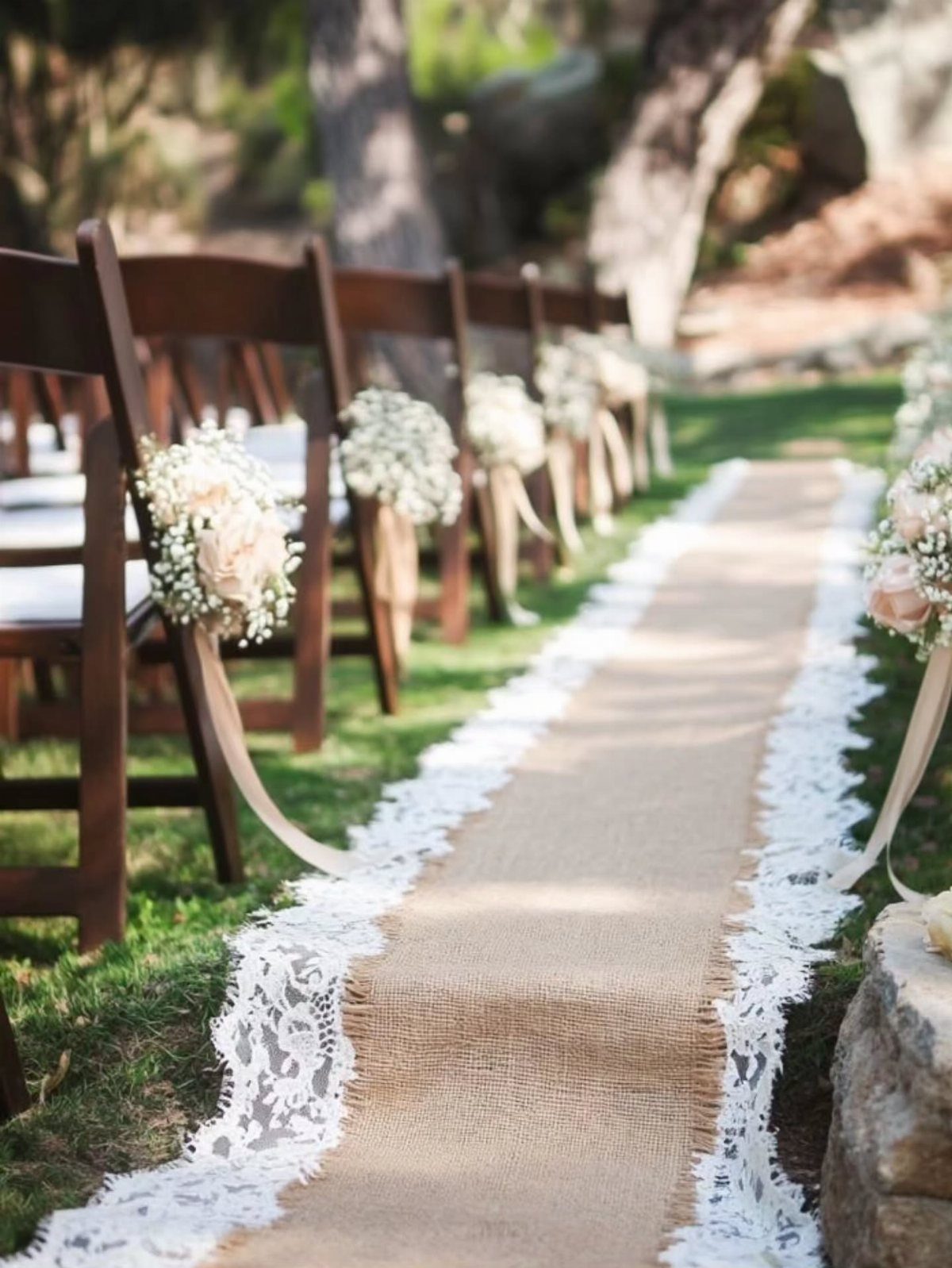 Burlap aisle runner with white lace trim between wooden chairs at outdoor wedding