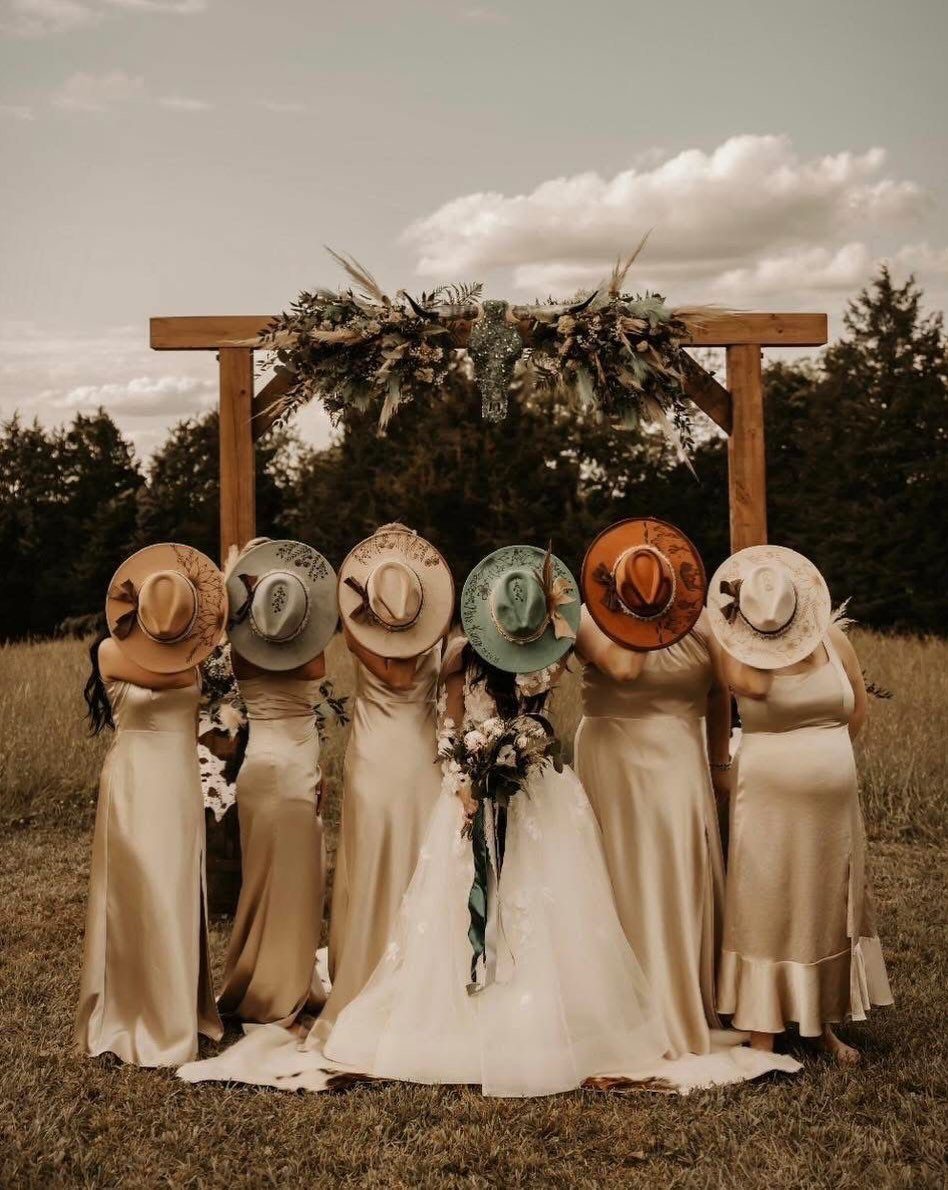 Bridesmaids in champagne dresses and wide-brimmed hats standing before a rustic wooden arbor.