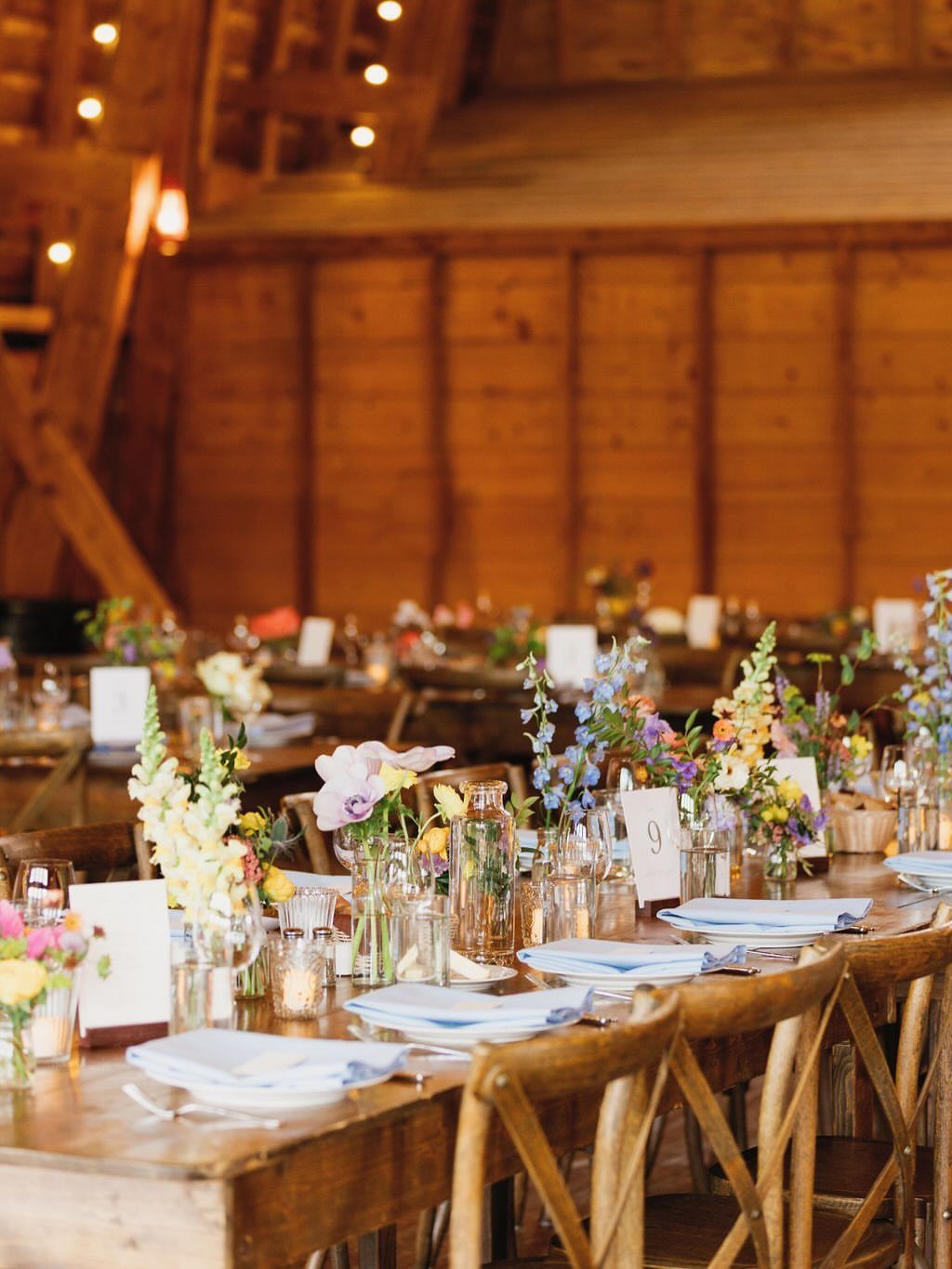 Colorful wildflower bouquets in glass vases on wooden farmhouse tables inside a rustic barn.