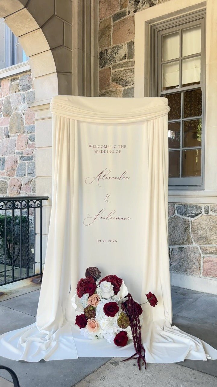 10 Wedding Venue Decorations for a Standout Celebration 5 White fabric wedding welcome sign with floral arrangement against a stone wall