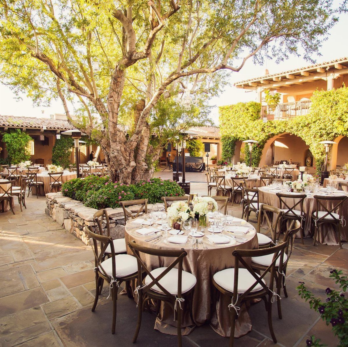 Wedding Venues Western Couples Love: 16 Ideas 2 Outdoor wedding reception with round tables on a stone patio under a large tree.