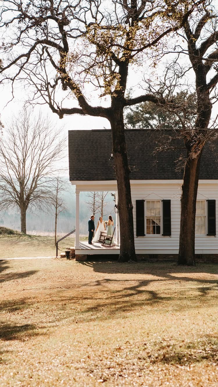 Wedding Venues Western Couples Love: 16 Ideas 15 White farmhouse with a bride and groom on the porch under large trees.